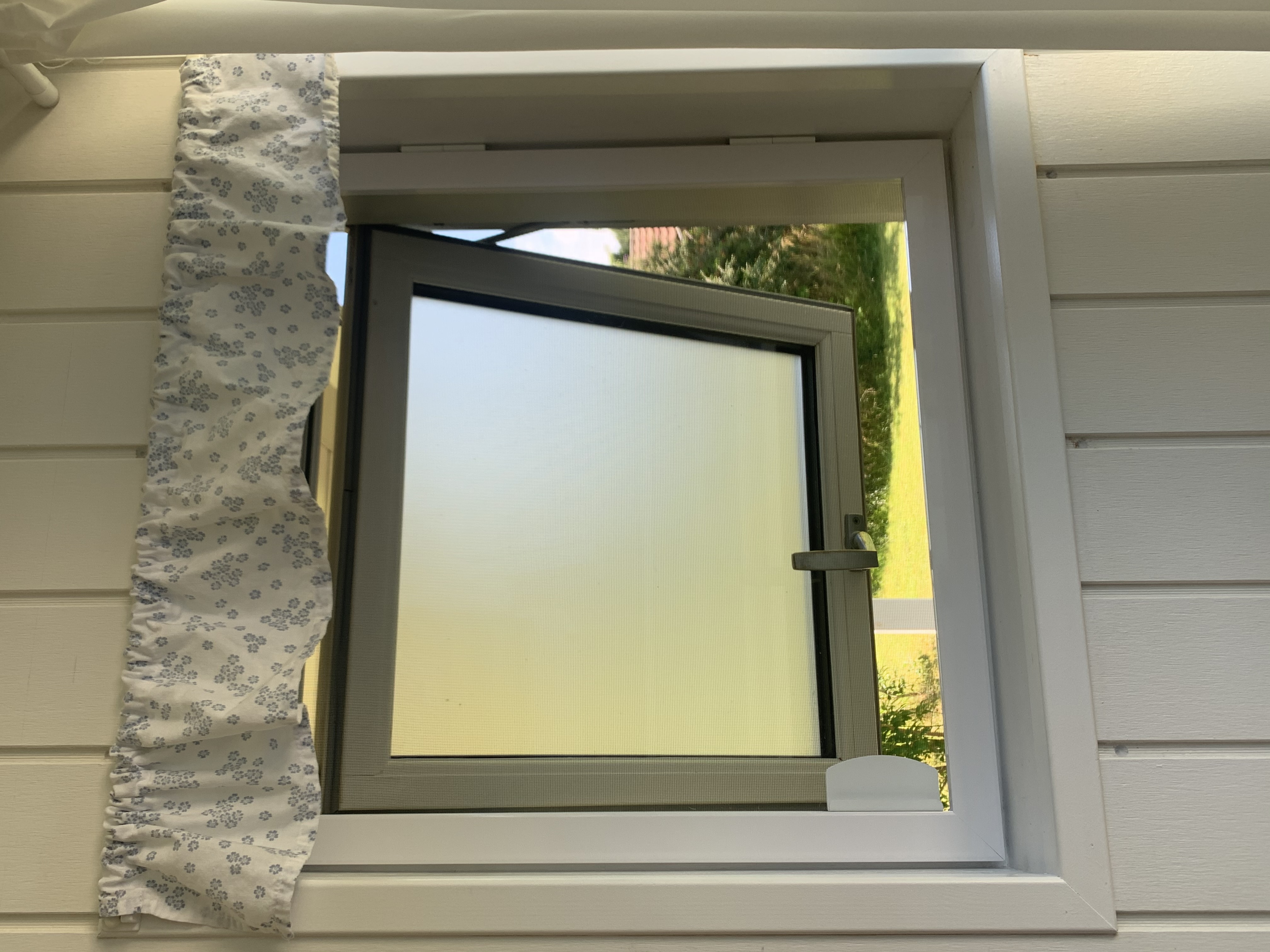 Ein weißes Fenster ist gekippt und von Innen sitzt ein weißer Insektenschutzdrehrahmen davor, den man mit einer Griffplatte nach Innen öffnen kann. - Schmidt Insektenschutzrahmen GmbH