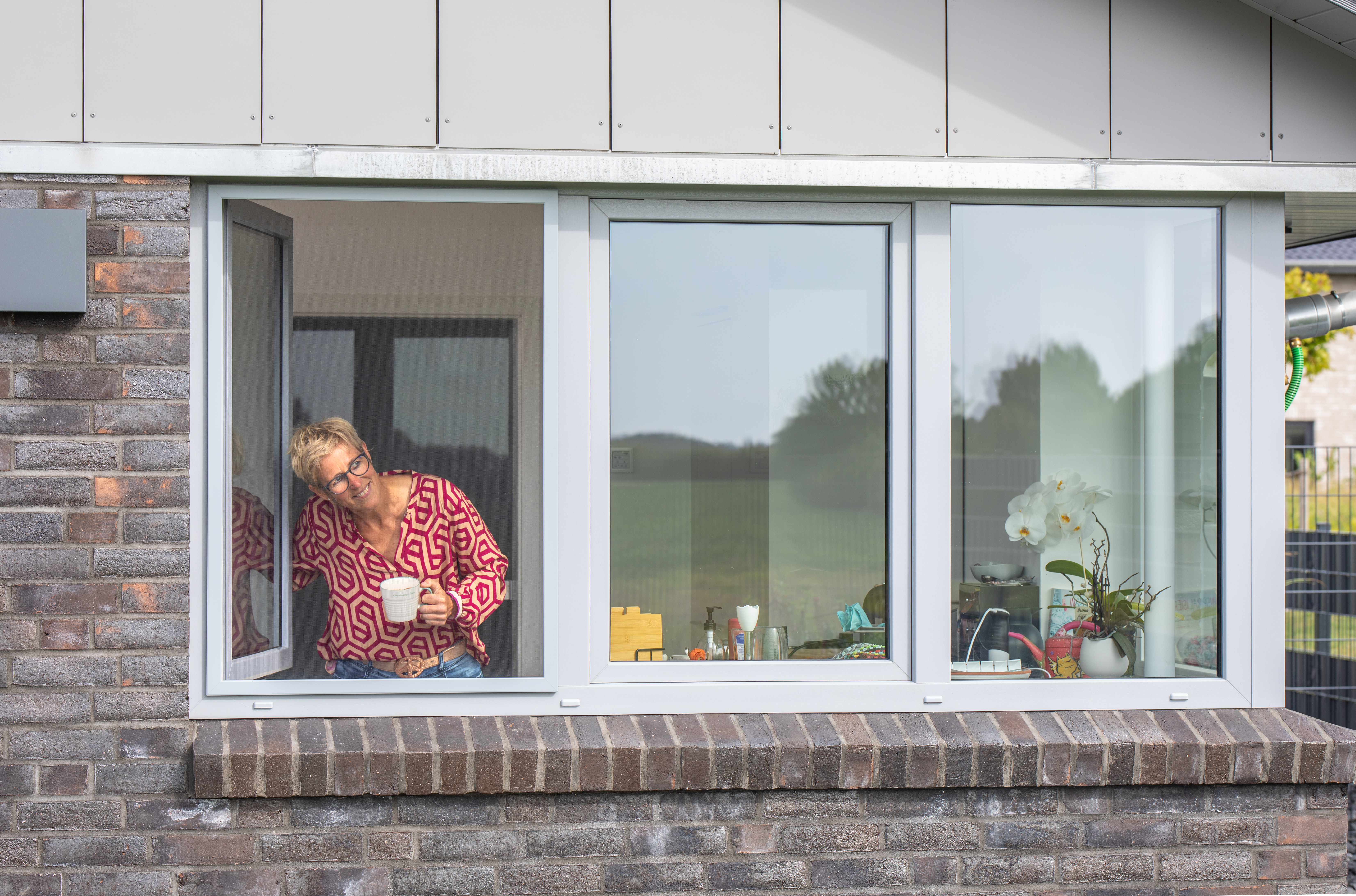 Frau mit Kaffeetasse in der Hand blickt durch geöffnetes Fenster mit Insektenschutz-Spannrahmen nach draußen