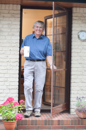Ein Mann geht mit einem Krug in der Hand durch eine braune Insektenschutzpendeltür. Diese Tür ist mit einer Sprosse und einem Sockelblech als Trittschutz ausgestattet. Rundherum sind blühende, bunte Blumen vor dem weißem Haus zu sehen. - Schmidt Insektenschutzrahmen GmbH